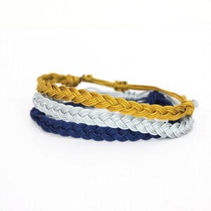 May include: Three braided cord bracelets in gold, silver, and navy blue. Each bracelet is adjustable with a sliding knot closure. The bracelets are stacked on top of each other, showcasing the different colors and textures.