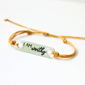 May include: A gold string bracelet with a silver charm that reads "I AM worthy".