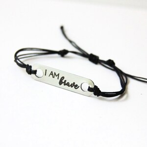 May include: A black string bracelet with a silver charm that says "I AM brave".