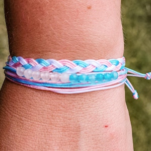May include: A multi-strand bracelet featuring a braided section in light blue, pink, and white cord. Below the braid, a single strand holds a row of small, round, translucent white beads followed by a row of small, round, translucent blue beads.