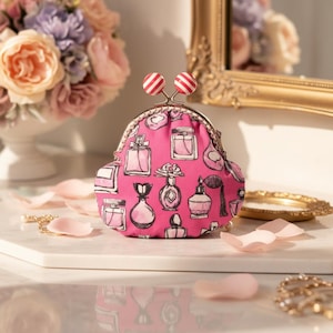 May include: A pink coin purse with a clasp closure, patterned with black and white perfume bottles. The purse has a decorative metal frame and is topped with two red and white striped spheres.
