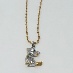 May include: A gold-toned chain necklace with a silver and gold-toned cat pendant. The cat pendant has green eyes and is sitting down. The necklace has a tag attached to it.