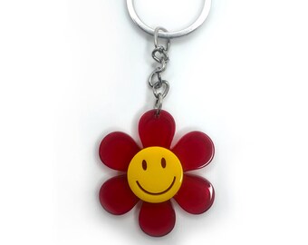 Peut inclure: Porte-clés en forme de fleur rouge avec un smiley jaune au centre. Le porte-clés a un anneau et une chaîne en métal argenté.