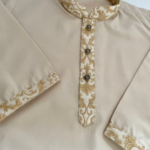 Puede incluir: Una camisa beige de manga larga con cuello mandar&iacute;n y ribetes bordados en dorado. La camisa tiene una abertura de botones y cinco botones. El texto "CHOTTI KOTI 3T" es visible en el cuello.