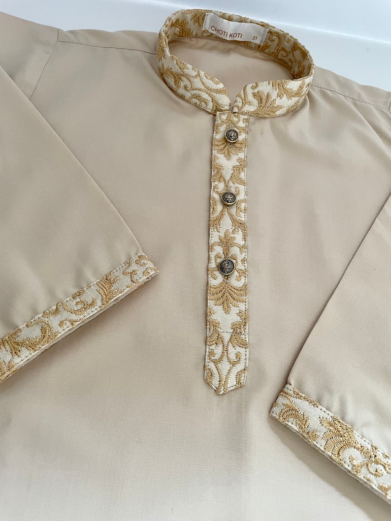 Puede incluir: Una camisa beige de manga larga con cuello mandar&iacute;n y ribetes bordados en dorado. La camisa tiene una abertura de botones y cinco botones. El texto "CHOTTI KOTI 3T" es visible en el cuello.