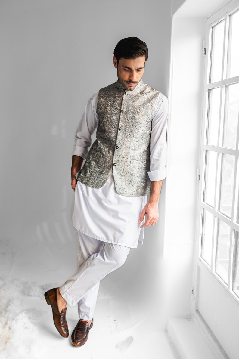 May include: A man wearing a white long-sleeved shirt with a silver and gray patterned vest. He is wearing white pants and brown leather shoes. He is standing next to a window.
