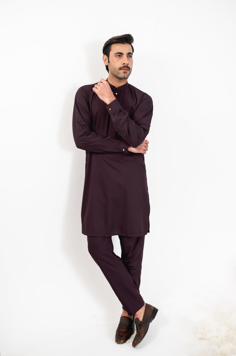 May include: A man wearing a long-sleeved, dark purple kurta with matching pants and brown leather shoes. He is standing with his arms crossed and looking to the side.