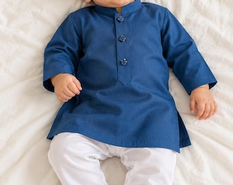 Puede incluir: Una túnica de manga larga en color azul oscuro con cuello mao y tres botones decorativos en la tapeta frontal, combinada con pantalones blancos lisos. La prenda presenta un diseño de color sólido con dobladillo recto y aberturas laterales.