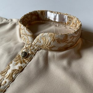 Puede incluir: Una camisa beige con un cuello bordado en oro. El cuello tiene un patr&oacute;n floral y la camisa tiene dos botones dorados.