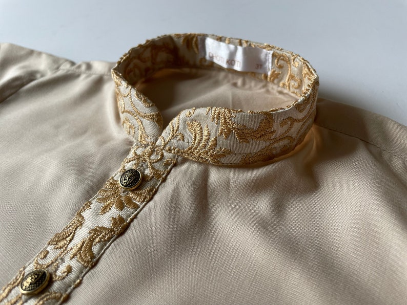 Puede incluir: Una camisa beige con un cuello bordado en oro. El cuello tiene un patr&oacute;n floral y la camisa tiene dos botones dorados.