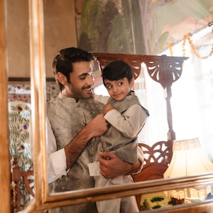 May include: A man and a young boy are reflected in a mirror. The man is wearing a gray patterned vest and a white shirt. The boy is wearing a white shirt and a gray patterned vest. They are smiling at each other.