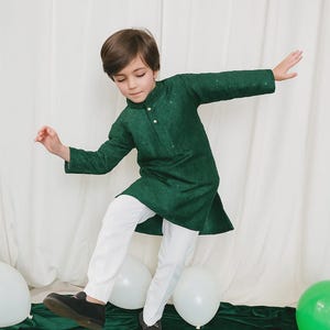 May include: A child's green kurta with white pants. The long-sleeved kurta has a mandarin collar and small embellishments. The white pants are straight-legged. The child is wearing black shoes. The background includes white curtains, green fabric, and balloons.