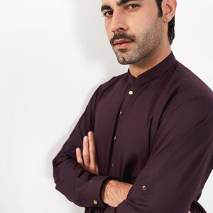 May include: A man wearing a burgundy long-sleeved shirt with a mandarin collar and gold buttons. He is standing with his arms crossed in front of a white background.