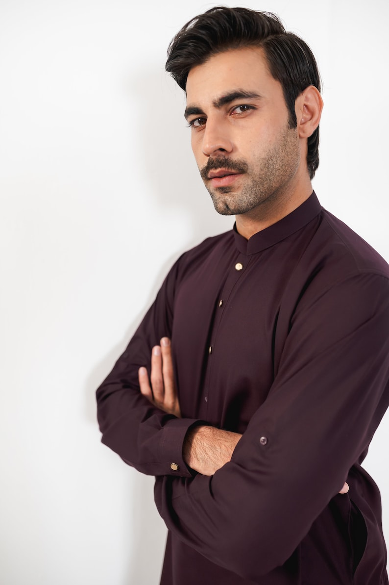 May include: A man wearing a burgundy long-sleeved shirt with a mandarin collar and gold buttons. He is standing with his arms crossed in front of a white background.