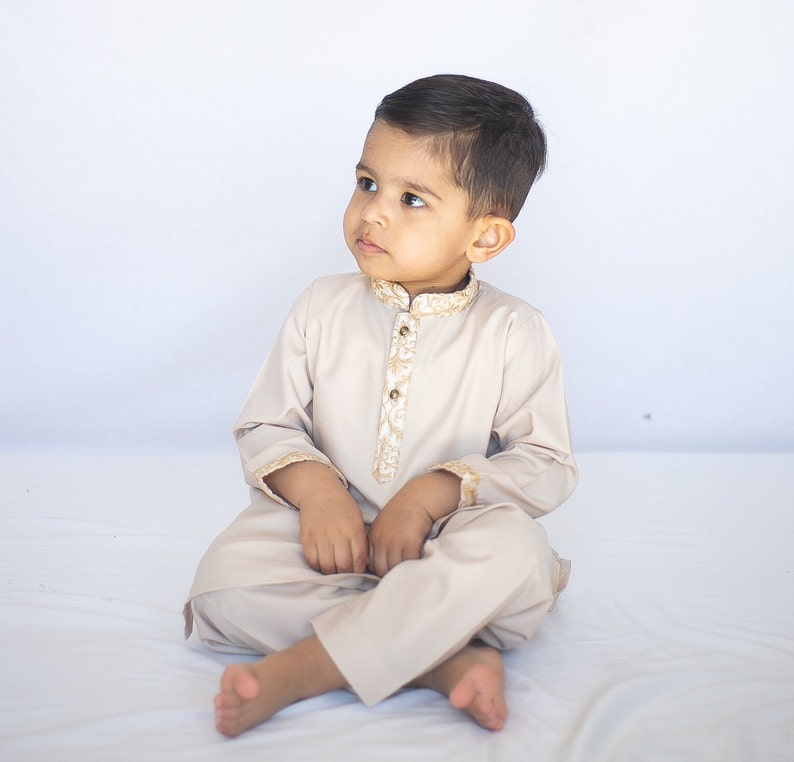Puede incluir: Un ni&ntilde;o peque&ntilde;o vistiendo un shalwar kameez beige con bordados dorados. Est&aacute; sentado en una superficie blanca y mirando hacia la izquierda.