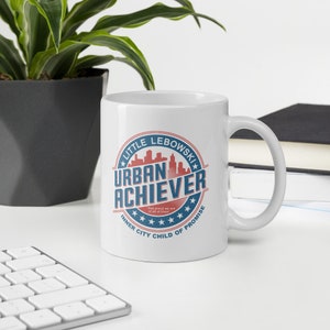 May include: White ceramic coffee mug with a blue and red circular design that says "Little Lebowski Urban Achiever Inner City Child of Promise".