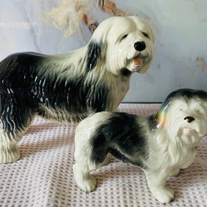 Puede incluir: Dos figuritas de cerámica de Old English Sheepdog. La figurita más grande está de pie con la cabeza inclinada hacia un lado. La figurita más pequeña está de pie con la cabeza mirando hacia adelante. Ambas figuritas están pintadas en tonos de blanco, negro y gris.