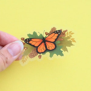 May include: A clear sticker with an orange and black butterfly on a bed of green leaves and brown acorns.