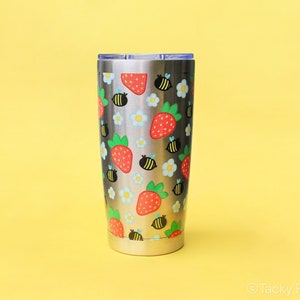 Puede incluir: Un vaso de acero inoxidable con un estampado repetido de fresas rojas, abejas negras y flores blancas y celeste. El vaso tiene una tapa plateada con un deslizador azul, sobre un fondo amarillo.