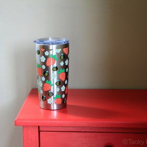 Puede incluir: Un vaso de acero inoxidable con tapa transparente, decorado con un dise&ntilde;o de fresas rojas, flores blancas y abejas negras y amarillas. El vaso es cil&iacute;ndrico y est&aacute; sobre una superficie roja.