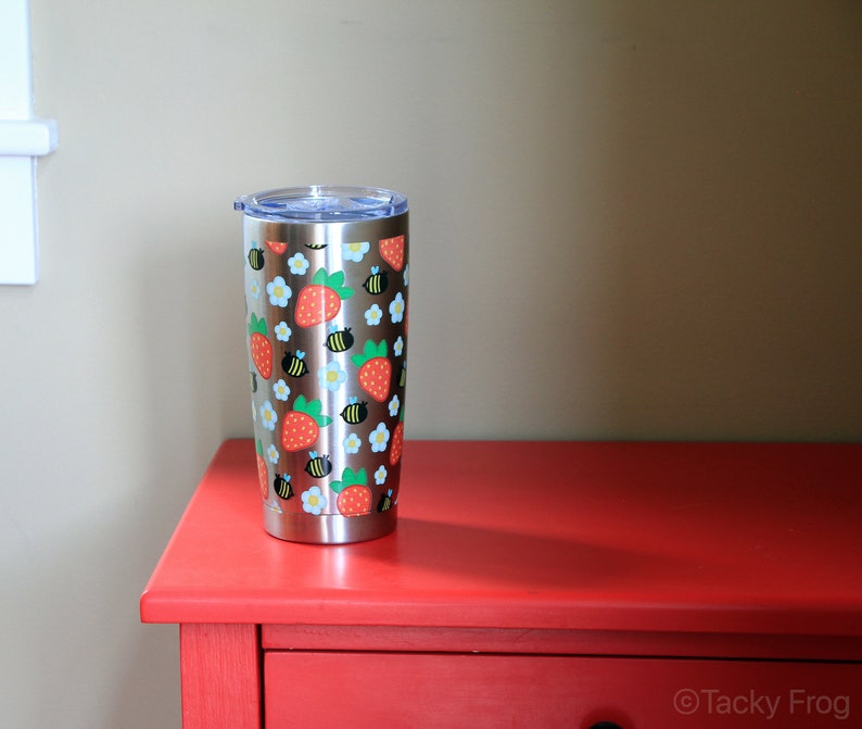 Puede incluir: Un vaso de acero inoxidable con tapa transparente, decorado con un dise&ntilde;o de fresas rojas, flores blancas y abejas negras y amarillas. El vaso es cil&iacute;ndrico y est&aacute; sobre una superficie roja.
