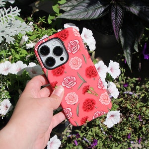 May include: A coral-colored phone case featuring a floral pattern of red and pink carnations and green leaves. The case has a black camera lens cutout and is held against a backdrop of greenery and white flowers.