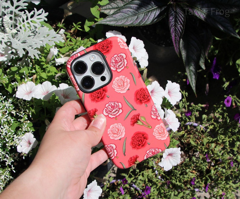 May include: A coral-colored phone case featuring a floral pattern of red and pink carnations and green leaves. The case has a black camera lens cutout and is held against a backdrop of greenery and white flowers.