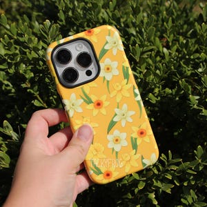 May include: Yellow phone case with a floral daffodil pattern. The case features white, yellow, and orange flowers with green leaves. The brand name "TACKYFROG" is printed on the bottom of the case.