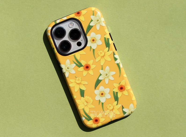 May include: Yellow phone case with a floral pattern of white and yellow daffodils and green leaves. The case has a black camera lens area and the text "TACKYFROG" at the bottom.