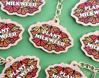 May include: Wooden keychains with a butterfly design in shades of orange, yellow, and black. The keychains have the text "PLANT MILKWEED" in white. Each keychain has a gold-colored chain and a wooden tag with a unique shape.
