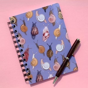 May include: A purple spiral bound notebook with a repeating pattern of colorful snails. The notebook is lying on a pink surface with a brown pen next to it.