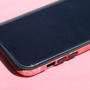 May include: A smartphone in a floral print case with a black border. The case features a pattern of pink and white flowers. The phone's screen is black, and the case has black buttons on the side.