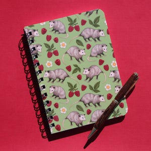 May include: A green spiral bound notebook with a repeating pattern of opossums and red raspberries. The notebook is lying on a red surface with a brown pen next to it.