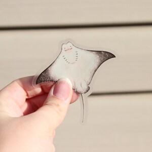 Peut inclure: Un autocollant transparent repr&eacute;sentant une raie manta souriante de dessin anim&eacute;. La raie a un corps blanc avec des pointes d'ailes grises et une longue queue. L'autocollant est tenu par une main sur un fond flou.
