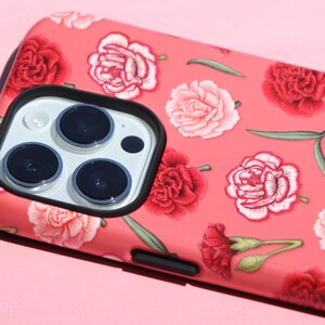 May include: A coral-colored phone case featuring a floral pattern of red and pink carnations and green leaves. The case has a black border around the camera lenses and a black button on the side. The phone case is on a pink background.
