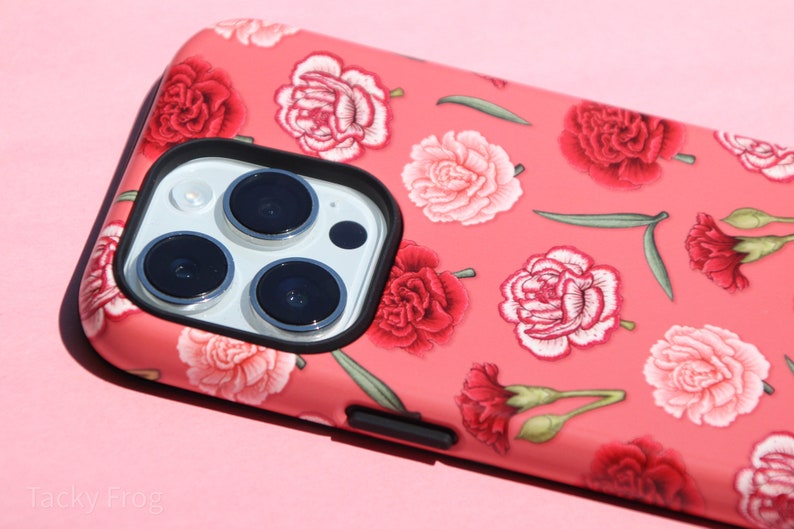 May include: A coral-colored phone case featuring a floral pattern of red and pink carnations and green leaves. The case has a black border around the camera lenses and a black button on the side. The phone case is on a pink background.