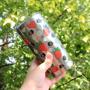 Puede incluir: Un vaso de acero inoxidable con un patr&oacute;n repetido de fresas rojas, abejas negras y amarillas y flores blancas y azules. El vaso se sostiene contra un fondo de follaje verde.