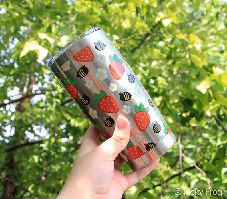 Puede incluir: Un vaso de acero inoxidable con un patr&oacute;n repetido de fresas rojas, abejas negras y amarillas y flores blancas y azules. El vaso se sostiene contra un fondo de follaje verde.