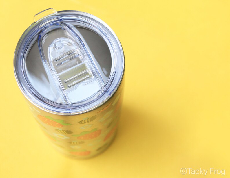 Puede incluir: Vaso de acero inoxidable con tapa de pl&aacute;stico transparente. El vaso presenta un patr&oacute;n repetido de fresas naranjas, hojas verdes y abejas amarillas. El vaso est&aacute; sobre un fondo amarillo brillante.
