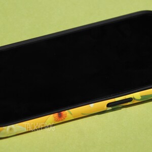 May include: A black smartphone with a yellow floral patterned case. The case features a design of orange and white flowers with green leaves. The phone screen is black and the case has the text "TACKYFROG".