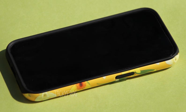May include: A black smartphone with a yellow floral patterned case. The case features a design of orange and white flowers with green leaves. The phone screen is black and the case has the text "TACKYFROG".