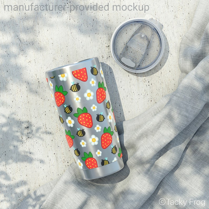 Puede incluir: Un vaso de acero inoxidable con tapa, con un estampado de fresas rojas, abejas amarillas y negras y flores blancas sobre un fondo gris. El vaso se muestra junto a su tapa y un pa&ntilde;o gris.