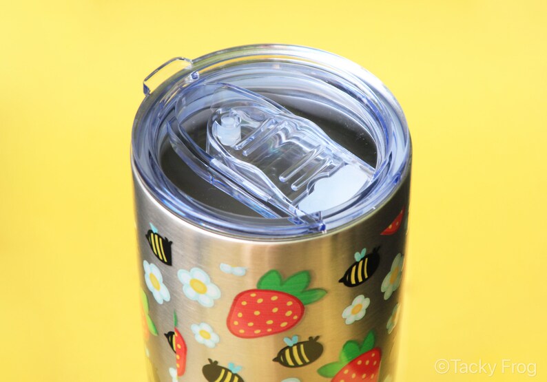 Puede incluir: Vaso de acero inoxidable con tapa de pl&aacute;stico transparente. El vaso est&aacute; decorado con ilustraciones coloridas de fresas, abejas y flores. La tapa tiene un cierre deslizante.