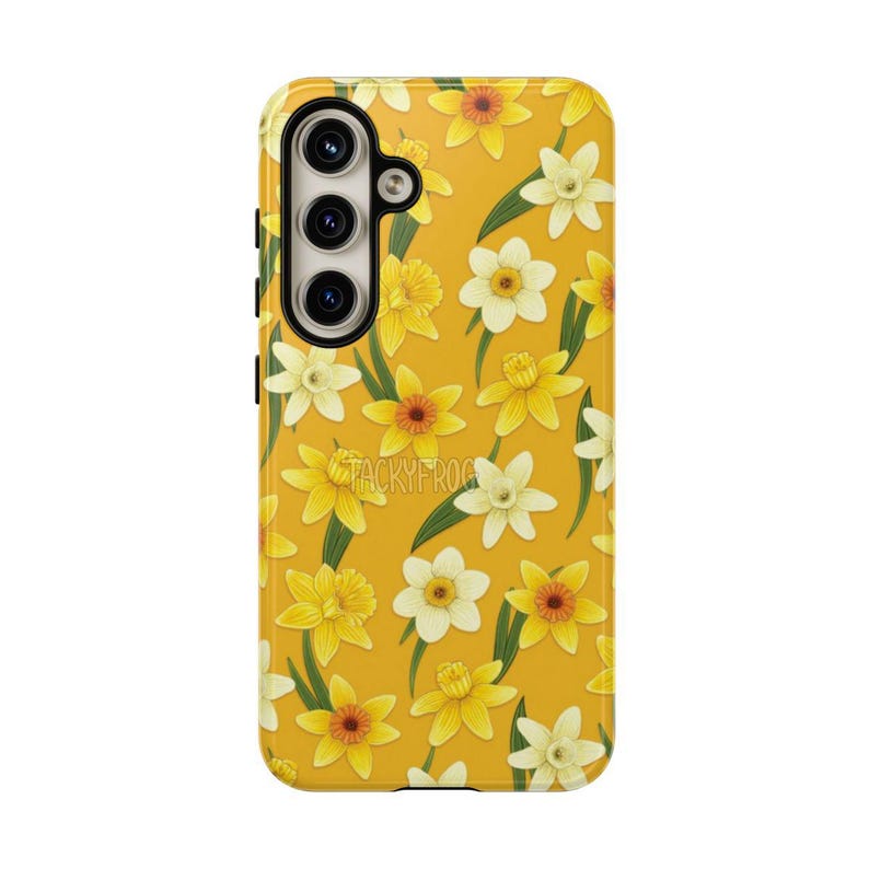 May include: A yellow phone case featuring a pattern of yellow and white daffodils with green leaves. The case has a camera cutout and the text "TACKYFROG" is visible. The case is designed to protect the phone while adding a floral aesthetic.
