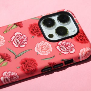 May include: A coral-colored phone case adorned with a floral pattern of red and pink carnations and green leaves. The case has a black camera lens frame and black side buttons. The phone case is designed to fit a smartphone.