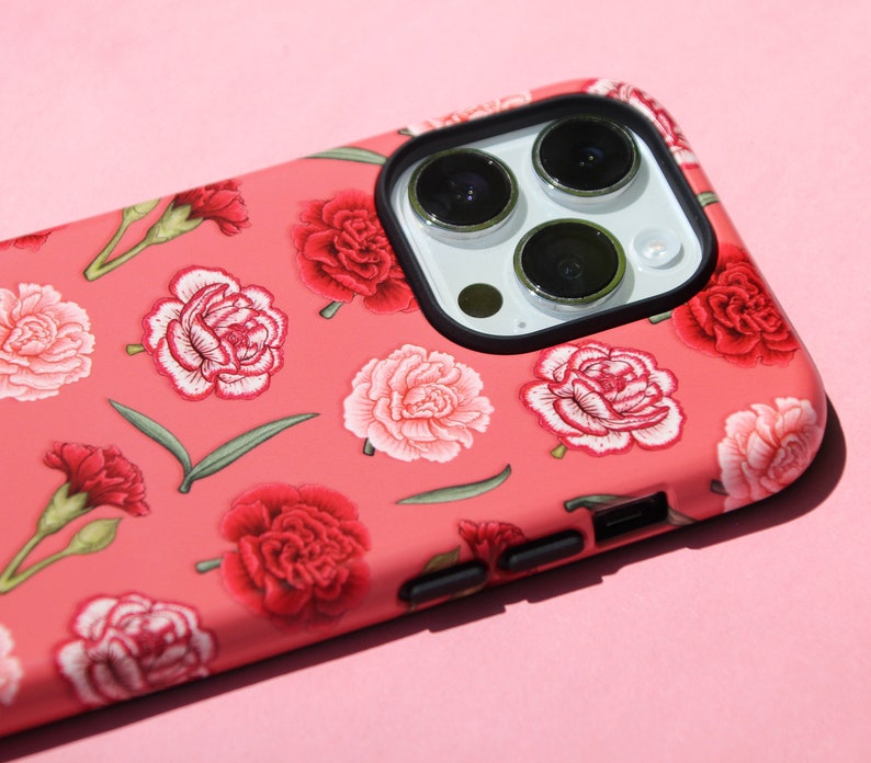 May include: A coral-colored phone case adorned with a floral pattern of red and pink carnations and green leaves. The case has a black camera lens frame and black side buttons. The phone case is designed to fit a smartphone.