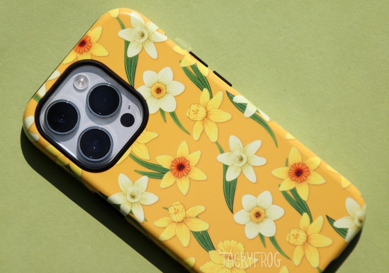 May include: Yellow phone case with a floral pattern of daffodils. The case features a repeating design of yellow and white flowers with green leaves. The phone's camera lenses are visible.