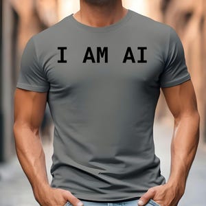 May include: Gray t-shirt with the text "I AM AI" in black block letters. The shirt is short-sleeved and appears to be made of a soft material. The person is wearing blue jeans.