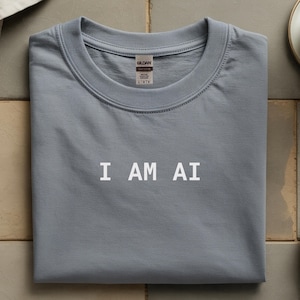 以下が含まれることがあります： 胸の中央に白い文字で「I AM AI」とプリントされた、スレートブルーのクルーネックTシャツ。襟元はリブ編みになっており、内側のネックラインにはGildanブランドのラベルが見え、このコットンシャツのサイズがLであることを示しています。