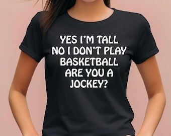 Puede incluir: Una camiseta negra de manga corta con texto en blanco que dice: YES I'M TALL NO I DON'T PLAY BASKETBALL ARE YOU A JOCKEY? La prenda tiene un cuello redondo y un corte estándar, mostrando el mensaje impreso claramente en el área del pecho.